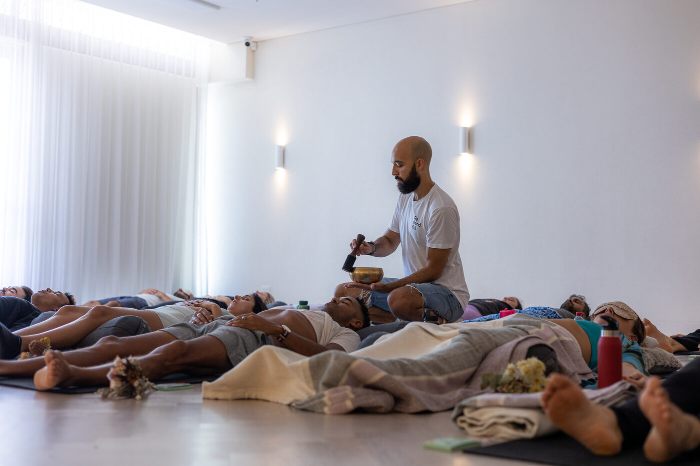 A room of participants lying down under blankets at the close of a Be Remindful group breathwork session, with Daniel Augustine seated at the front holding a singing bowl.